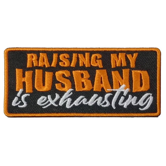 A rectangular embroidered patch with the text 'RAISING MY HUSBAND is exhausting' on a black background with orange and white embroidery.