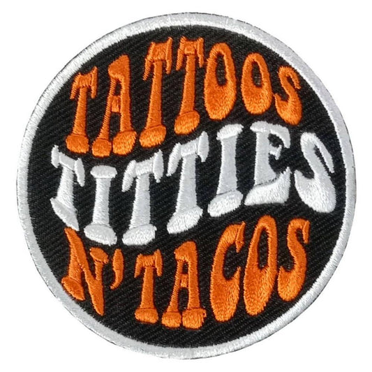 A circular embroidered patch with the text 'TATTOOS TITTIES N'TACOS' in bold white and orange letters on a black background.