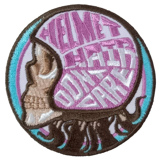 A circular embroidered patch featuring a skull design with text around it, predominantly in black, purple, and white colors.