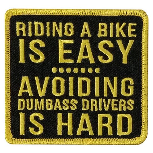 A black and yellow embroidered patch with the text 'RIDING A BIKE IS EASY AVOIDING DUMBASS DRIVERS IS HARD'.