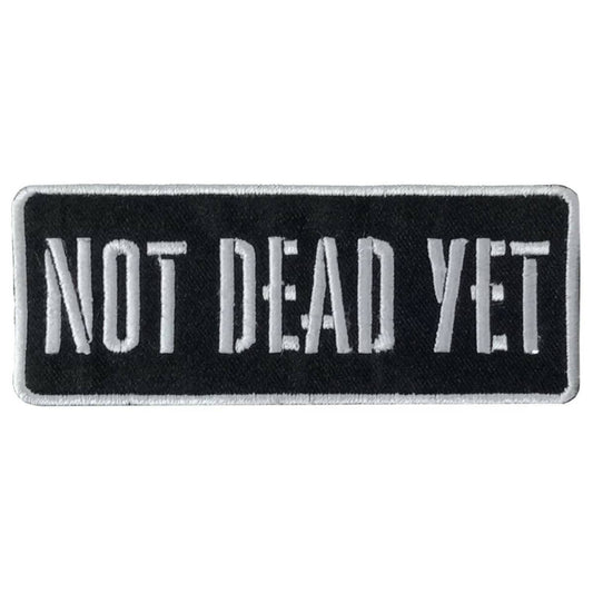 A black and white embroidered patch with the text 'NOT DEAD YET' in bold, capital letters on a contrasting white background.